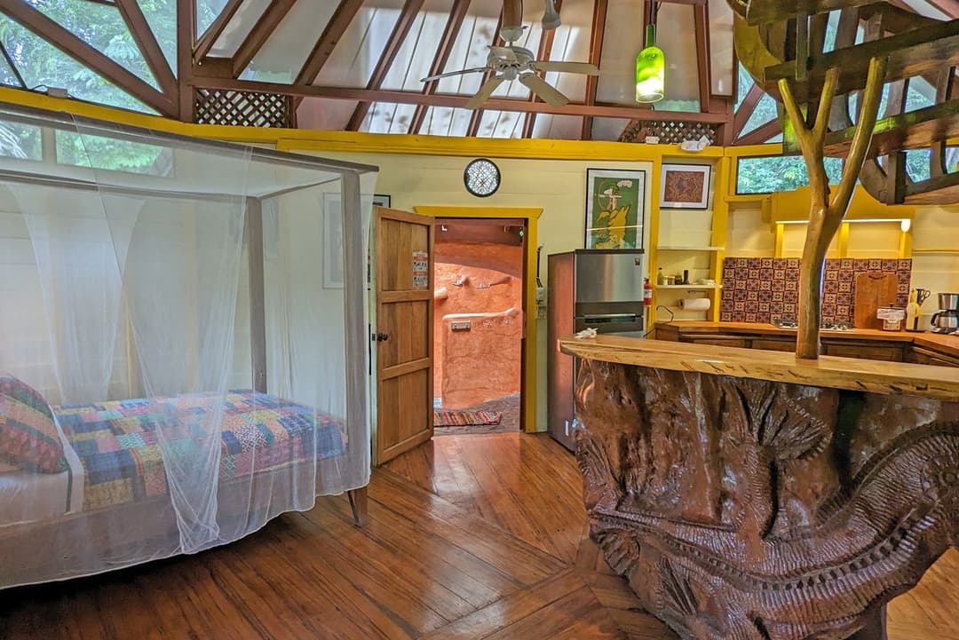 Open-plan ground floor — queen bed under mosquito net beside the kitchen with hand-carved Nispero bar