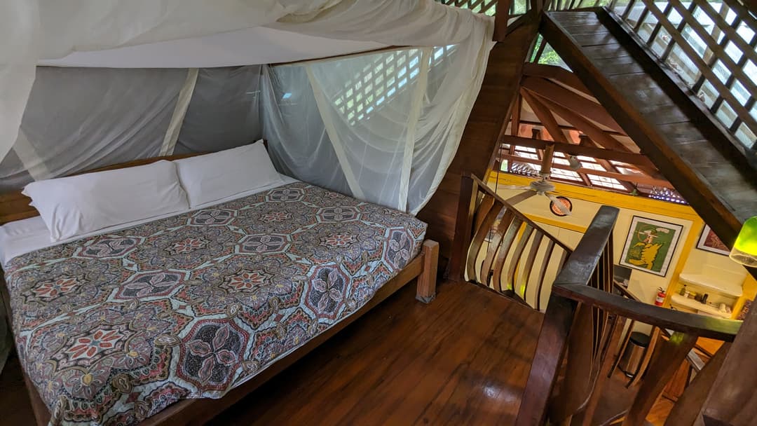 King bed upstairs dressed in a patterned cotton spread, mosquito net pulled back, ceiling fan and tropical artwork